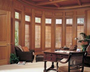 A study with wood-paneled curved walls, floor-to-ceiling windows with blinds, a dark wood desk, and wicker chairs.