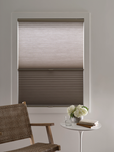 A window with top-down, bottom-up cellular shades in a beige room with a small side table holding flowers and a book.