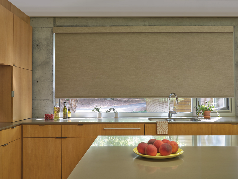 A kitchen with natural wood cabinets, a gray concrete wall, a wide window with a tan shade, and peaches on the island.