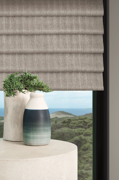 A textured, taupe Roman shade hangs over a window with a coastal view, beside two vases on a rounded pedestal.