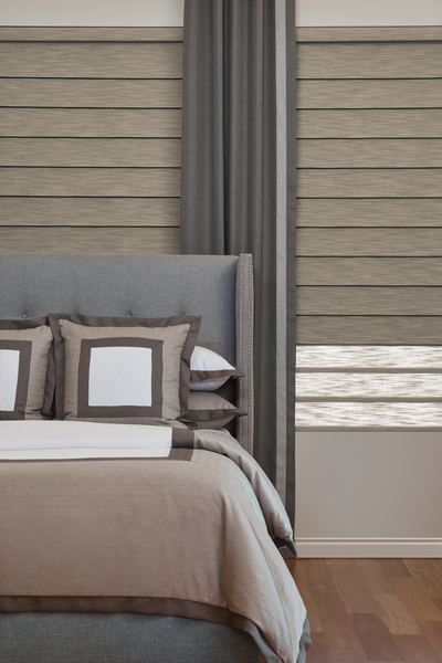 A bedroom featuring a grey upholstered headboard, tan bedding, and horizontal layered shades by a grey curtain.