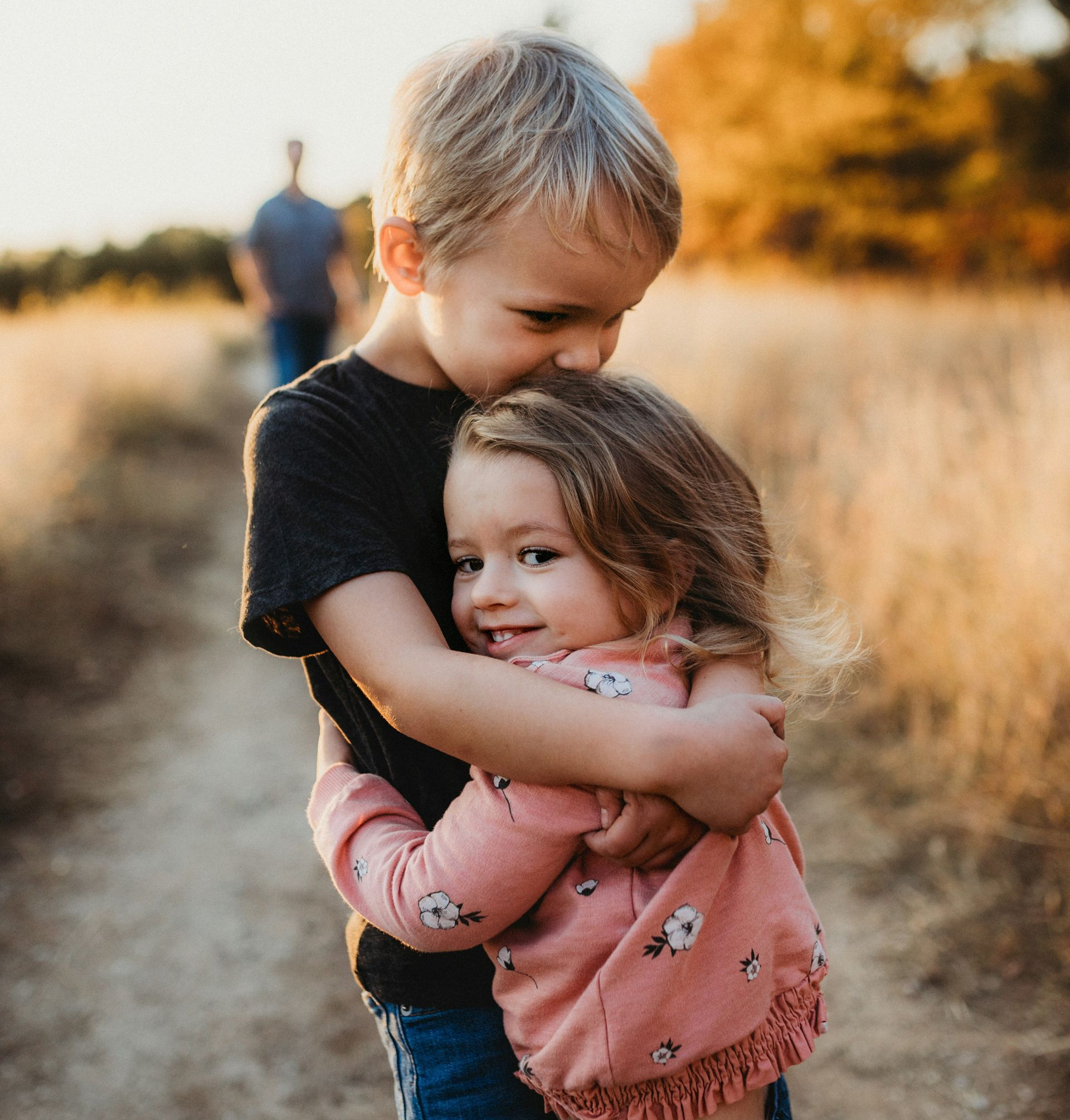 Boy hugging and kissing a girl on the head. They are outside on a dirt path with tall grass, warm sunlight.