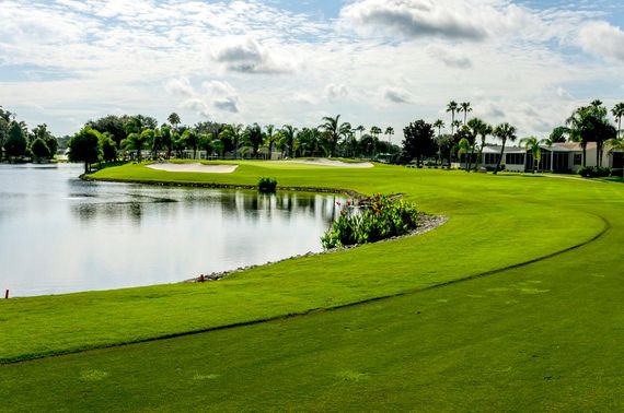 Lush green golf course with a lake, trees, and buildings under a cloudy sky.