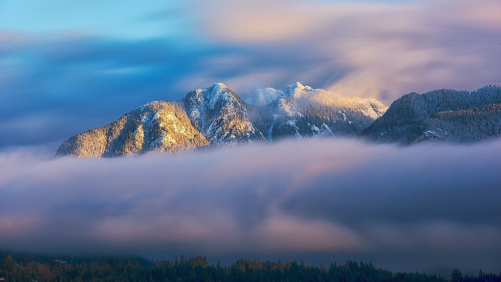 A mountain covered in snow and clouds with trees in the foreground.