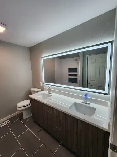 Modern bathroom with a large backlit mirror, dual sinks, and a dark tile floor.