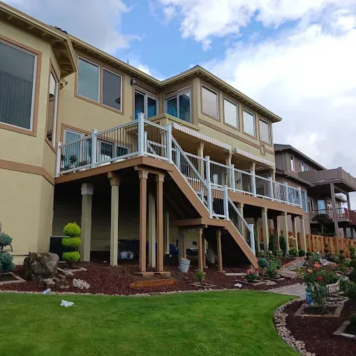 Two-story beige house with deck and stairs, glass railing, on a grassy lawn.