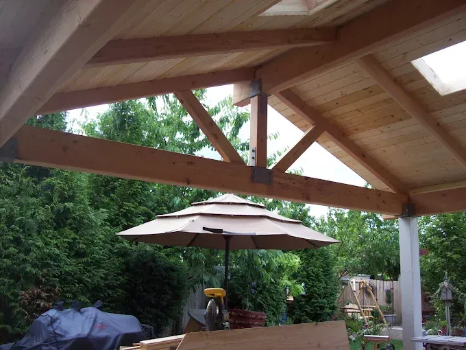 Wooden patio with a truss and skylight, looking out to a backyard with an umbrella and trees.