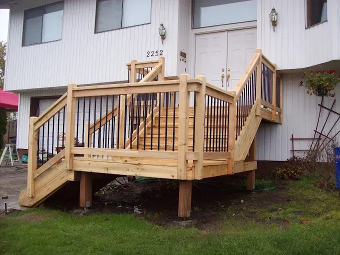 Wooden deck with stairs leading to a white house. Black railings, green grass.