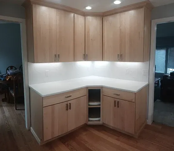 Light wood kitchen cabinets in a corner with a white countertop and under-cabinet lighting.