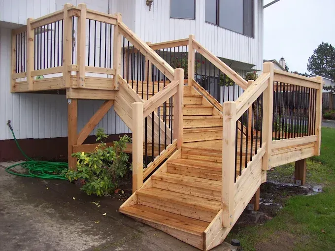 Wooden deck and staircase with black railing, built onto a white house with a green lawn.