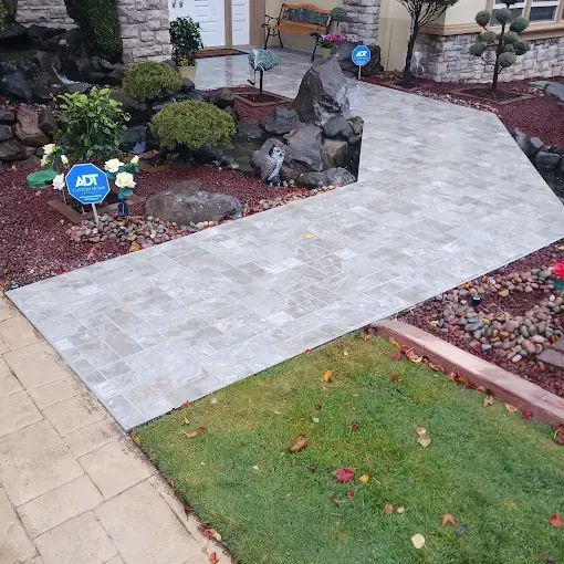 Paved walkway curves past a front yard with landscaping, including bushes and a small pond.