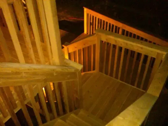 Wooden outdoor staircase with railings, steps leading down, well-lit with warm-toned light.