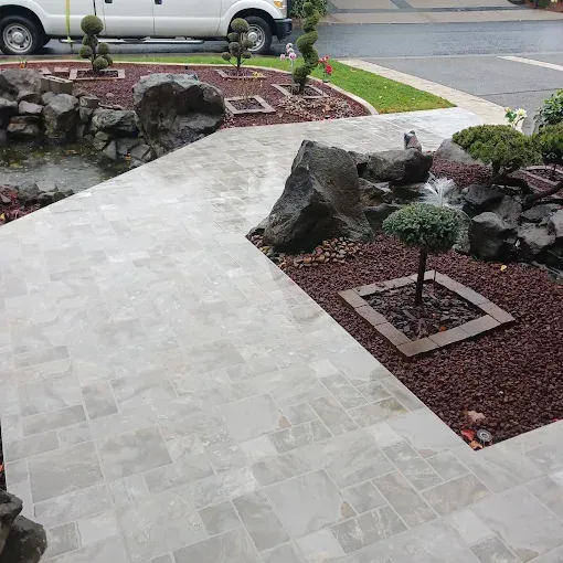 A light gray stone walkway leads to a landscaped yard with a small pond and bushes, near a road.