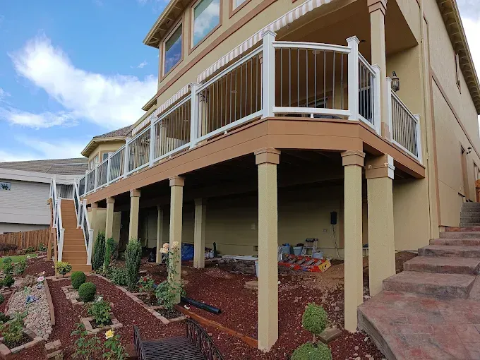 Two-story deck with glass railings. Tan siding, brown deck, white columns, landscaping, and blue sky.