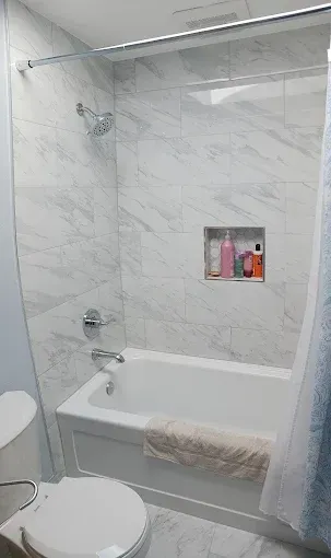 Bathroom with marble-like tiled shower, bathtub, toilet. Showerhead, built-in shelf with bottles, bath mat.