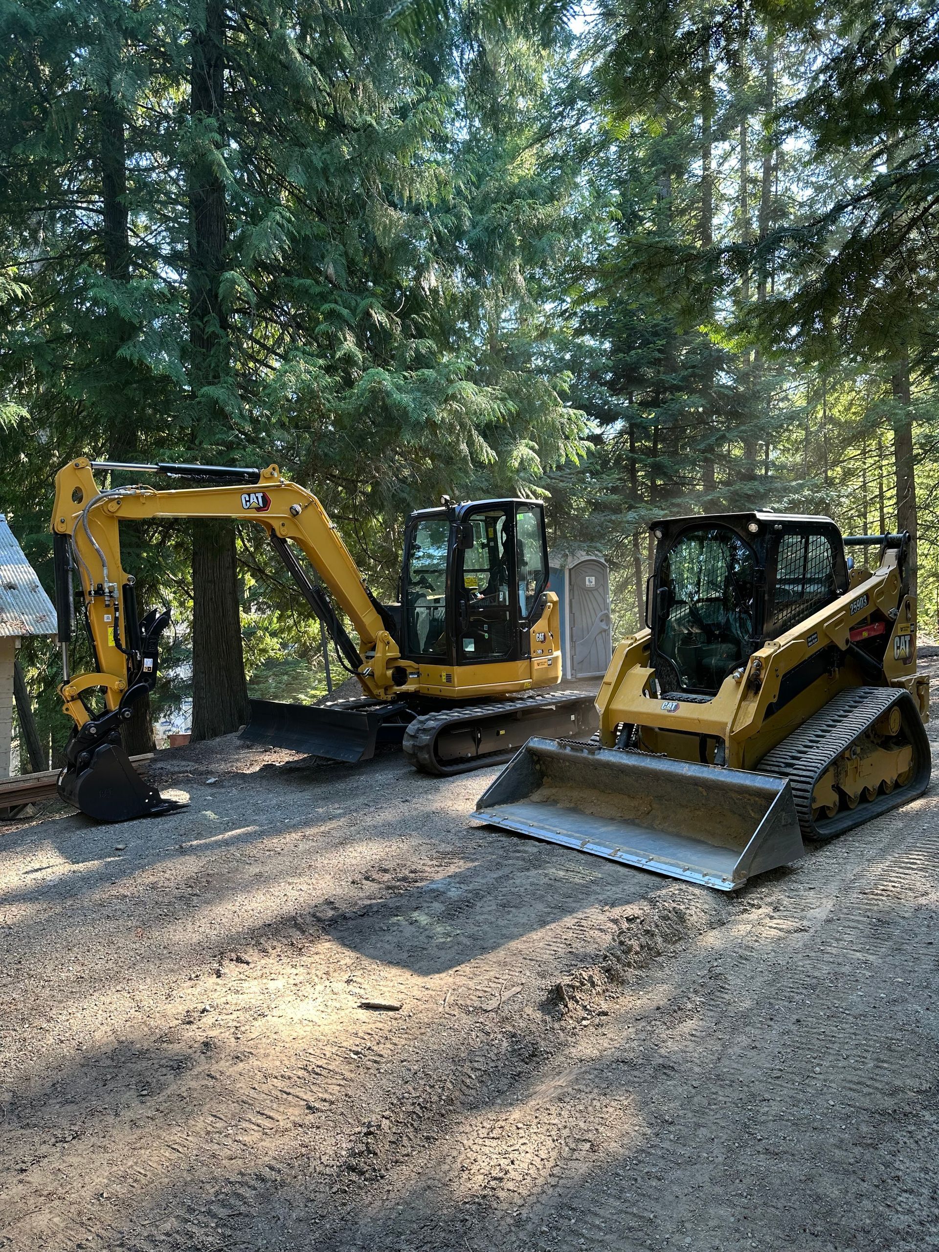 Yellow excavator and skid steer on a gravel surface, surrounded by trees.