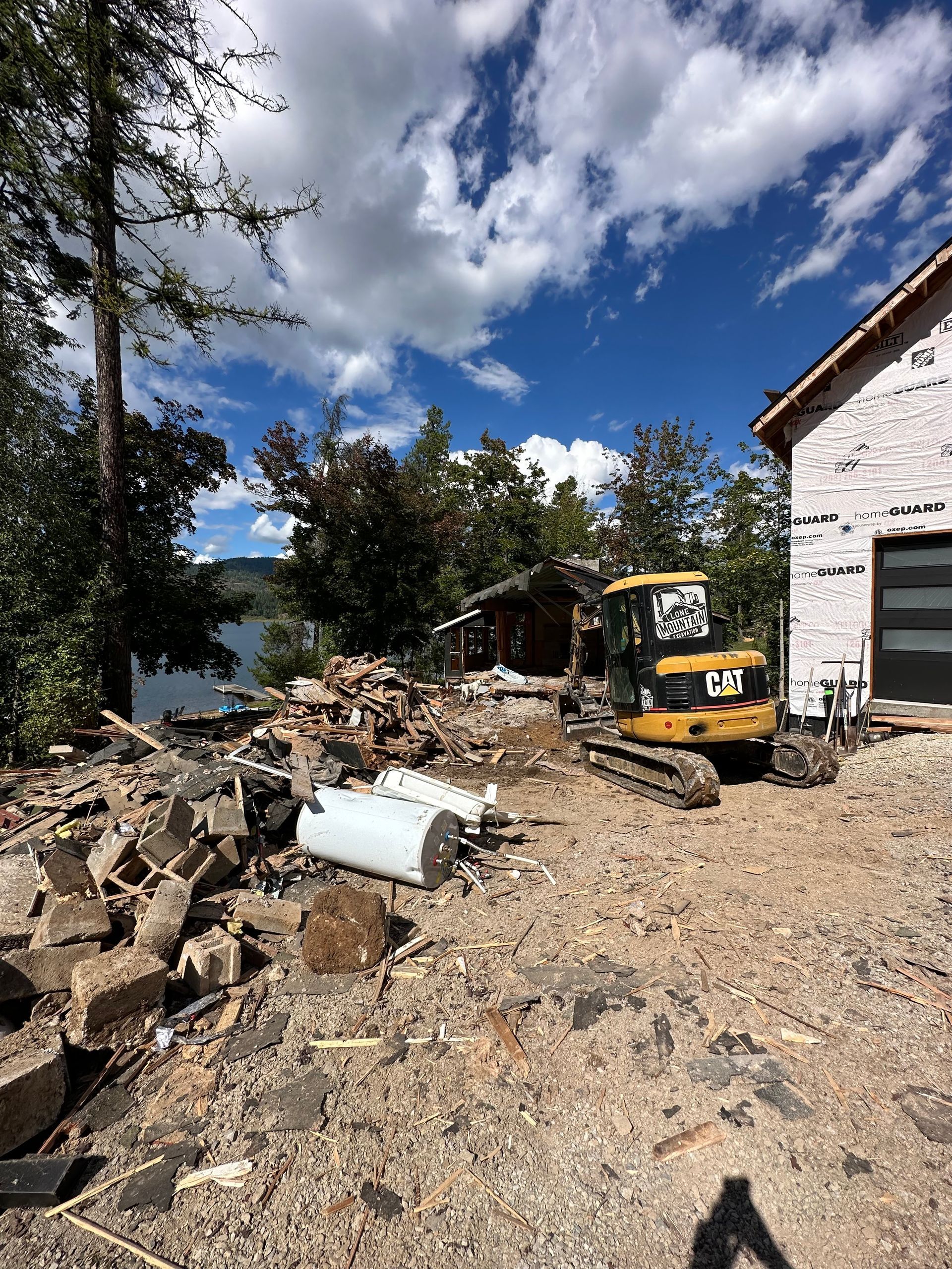 Construction site with a small excavator, demolition debris, a lake in the background, and a partially built house.
