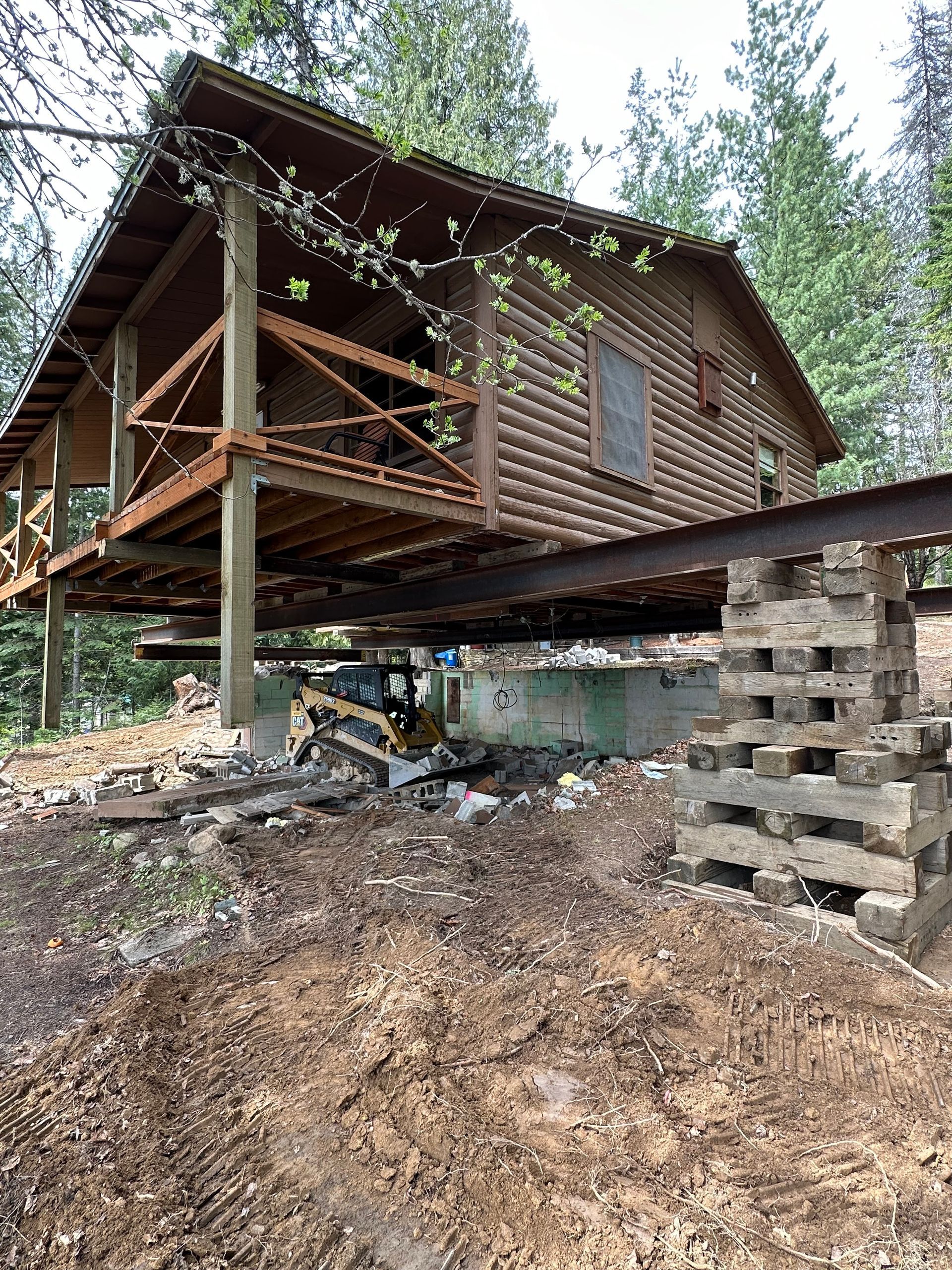 Log cabin raised on steel beams, supported by concrete and brick, in a wooded area. Construction in progress.