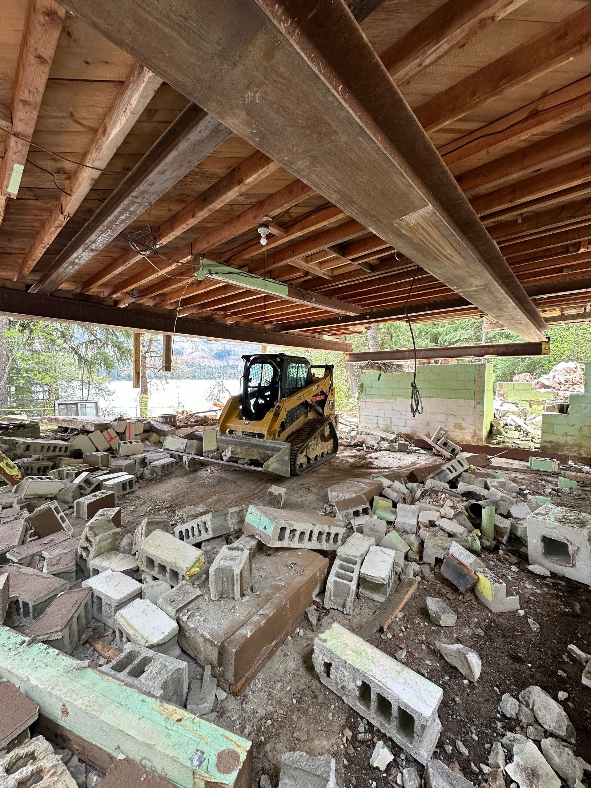 Demolition site with a small yellow skid steer, debris, and exposed overhead beams.