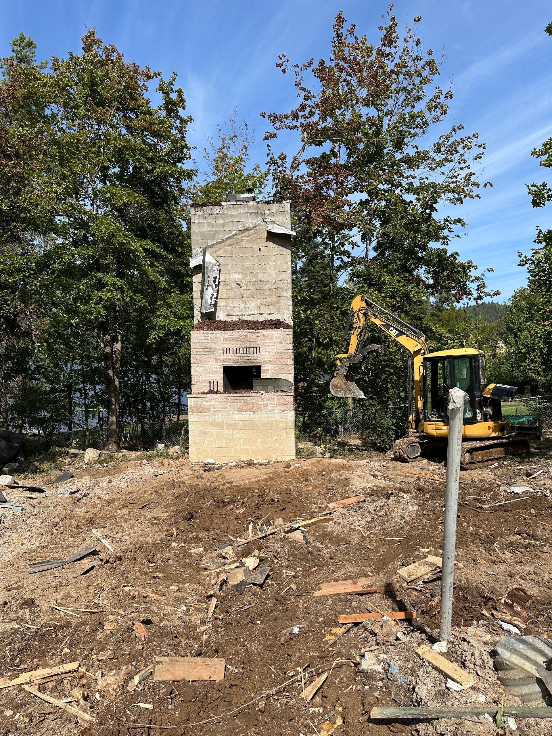 Brick chimney standing amid construction debris, with a yellow excavator in the background.
