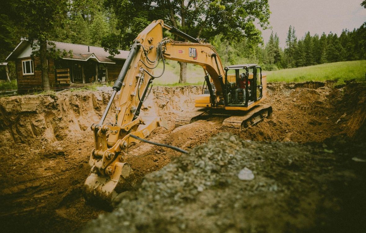 Yellow excavator digging a large pit in front of a weathered wooden cabin.