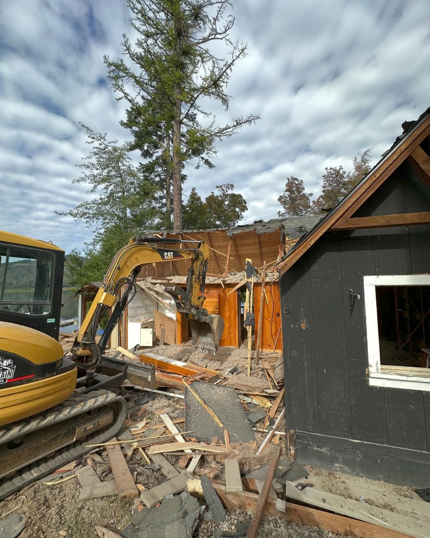 Demolition of a building. Yellow excavator tears down wooden structure; black house next to it.