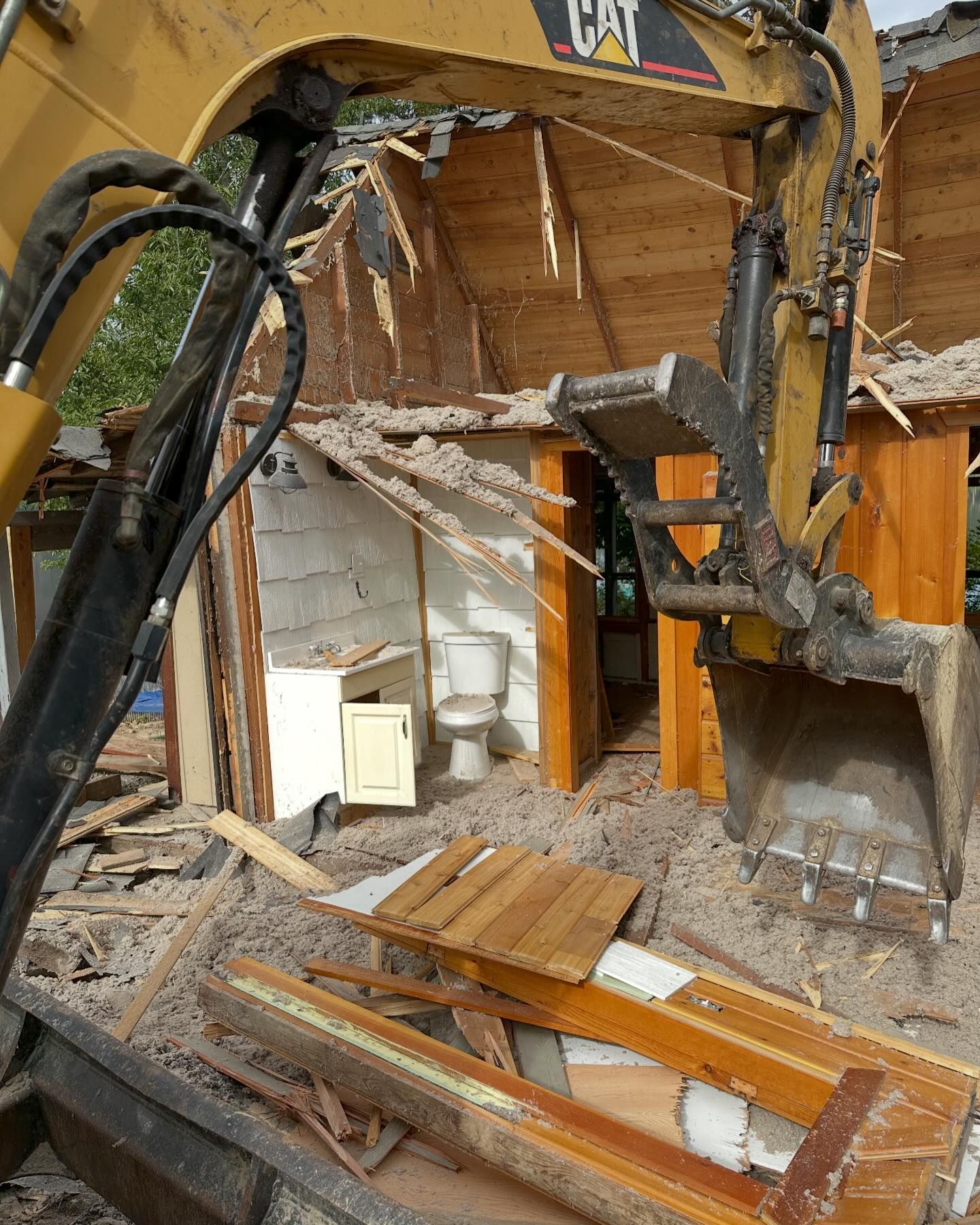 Excavator demolishing a small wooden building, exposing a toilet and interior.