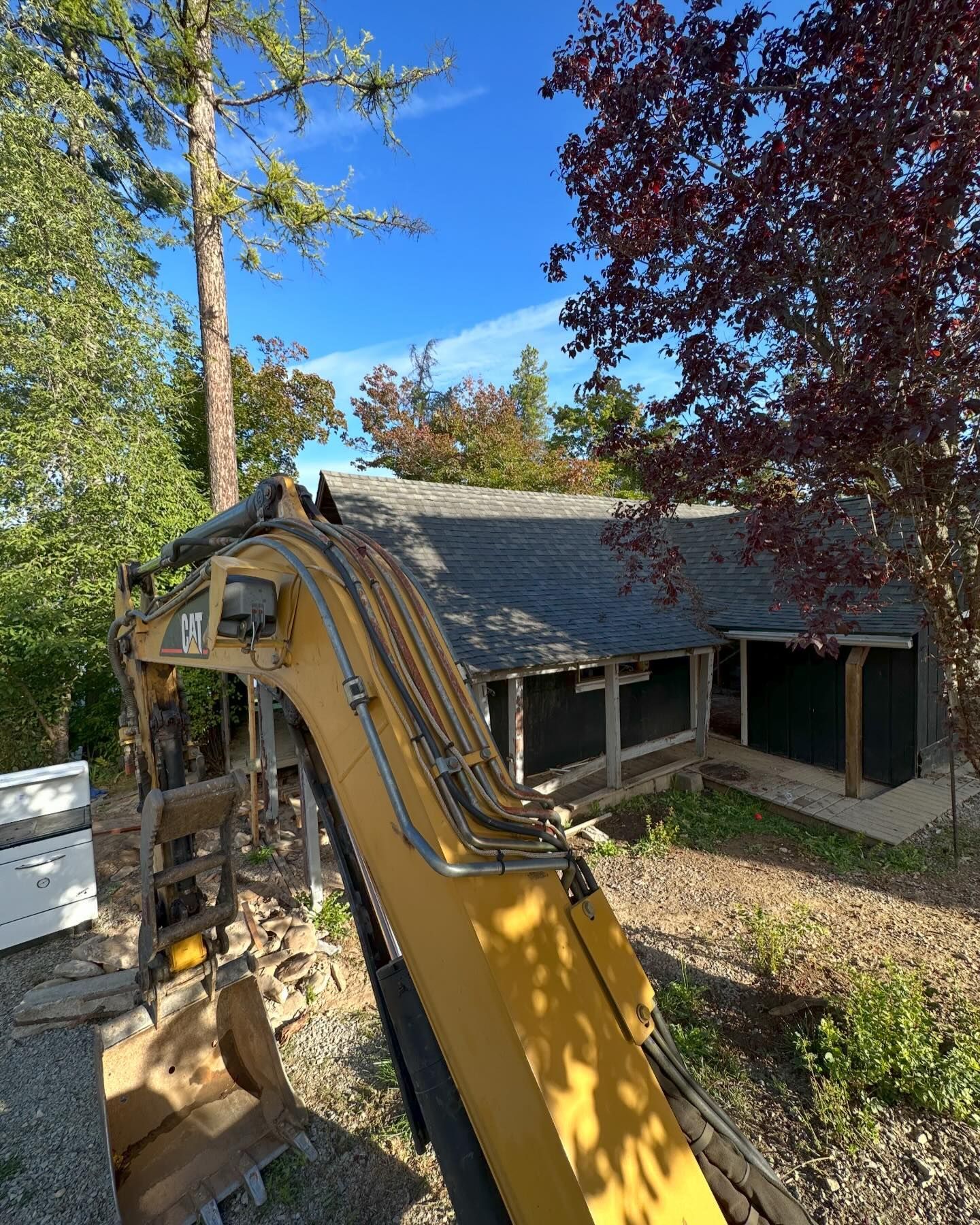 Yellow excavator working near a small building with a dark roof and surrounding trees under a blue sky.