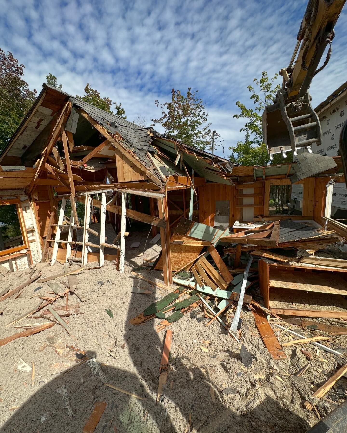 Demolition of a wood-framed house with excavator. Debris and exposed framing under blue sky with clouds.