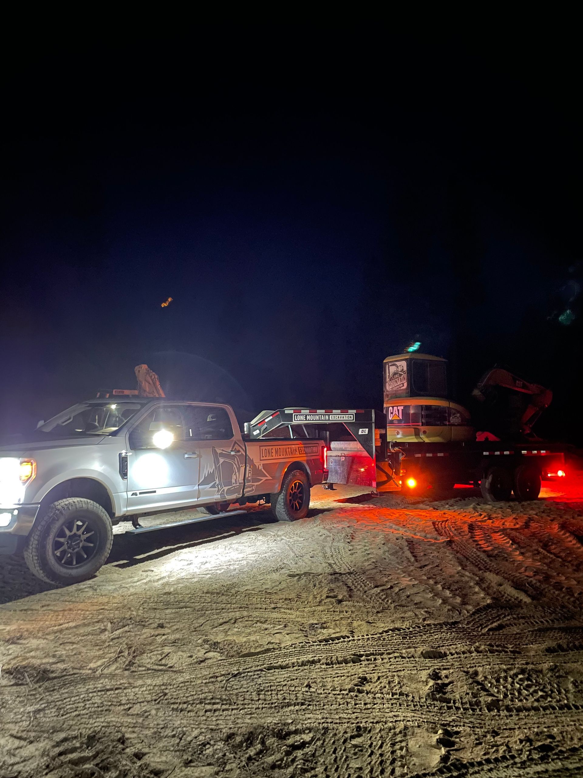 A pickup truck with headlights on towing a trailer with equipment at night on a dirt road.