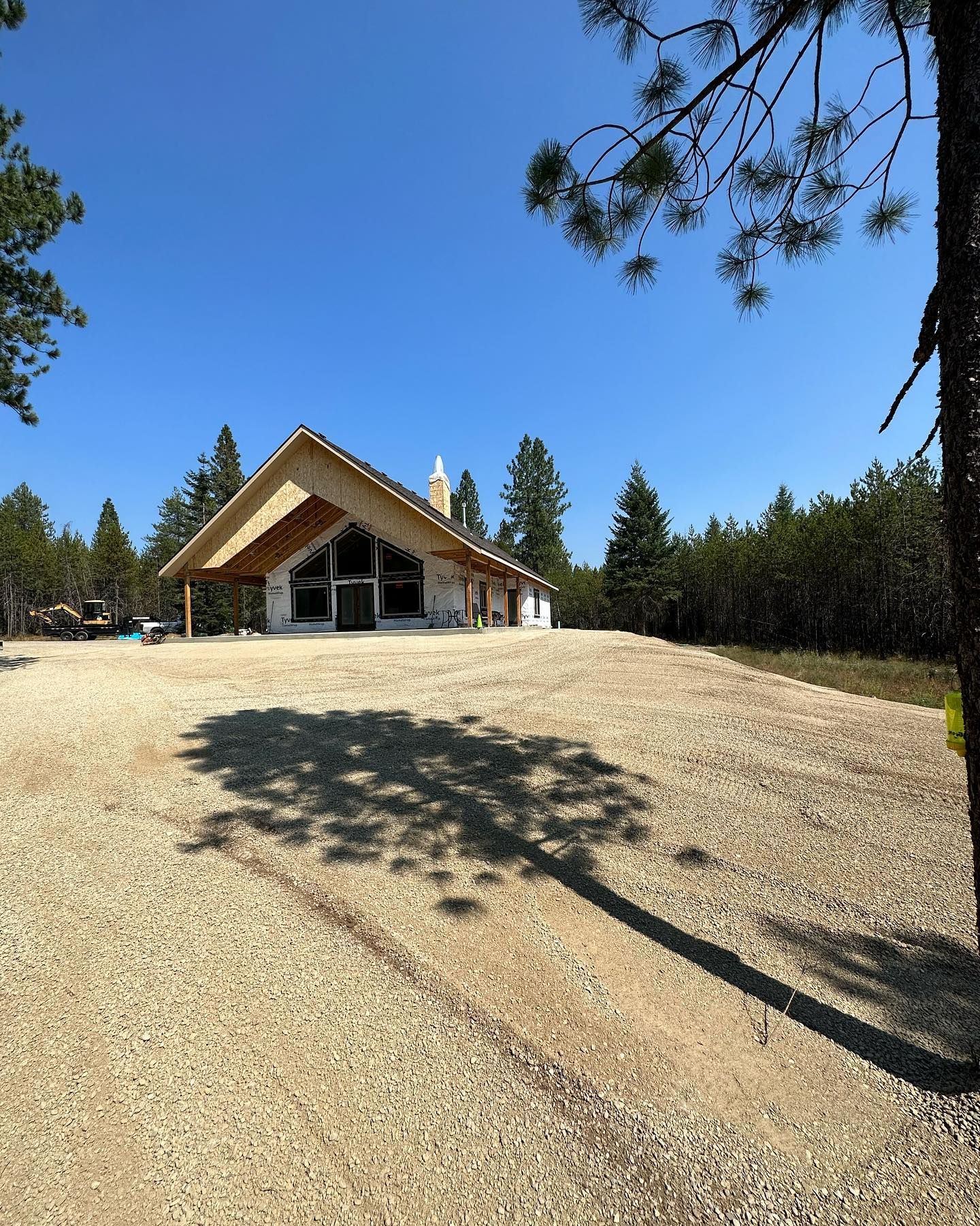 Building under construction with tall angled roof, set in a gravel lot surrounded by trees under a blue sky.