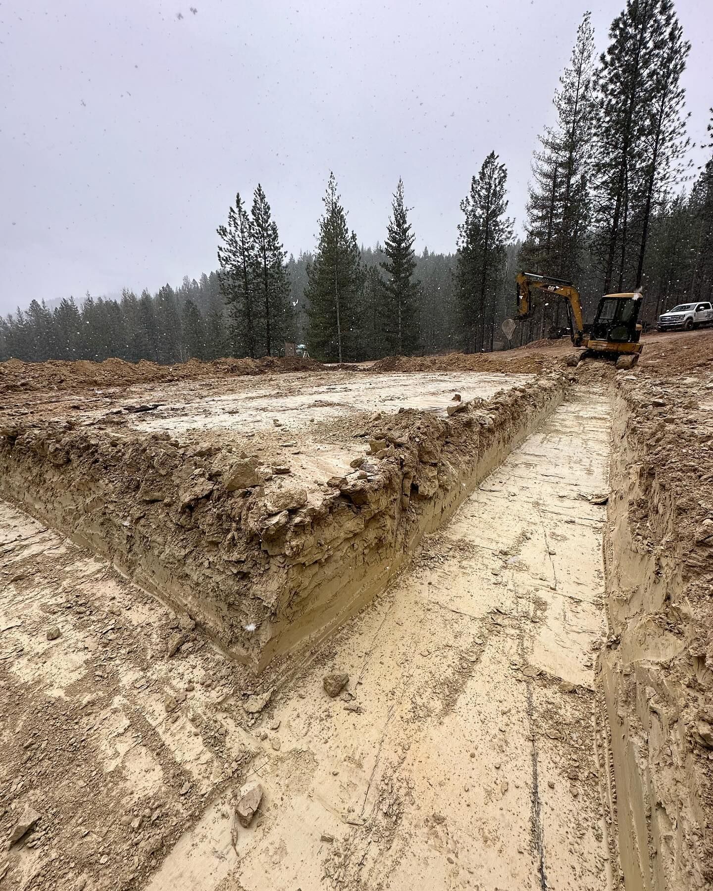 Construction site: Excavator digging foundation trenches in muddy ground during a snowfall.