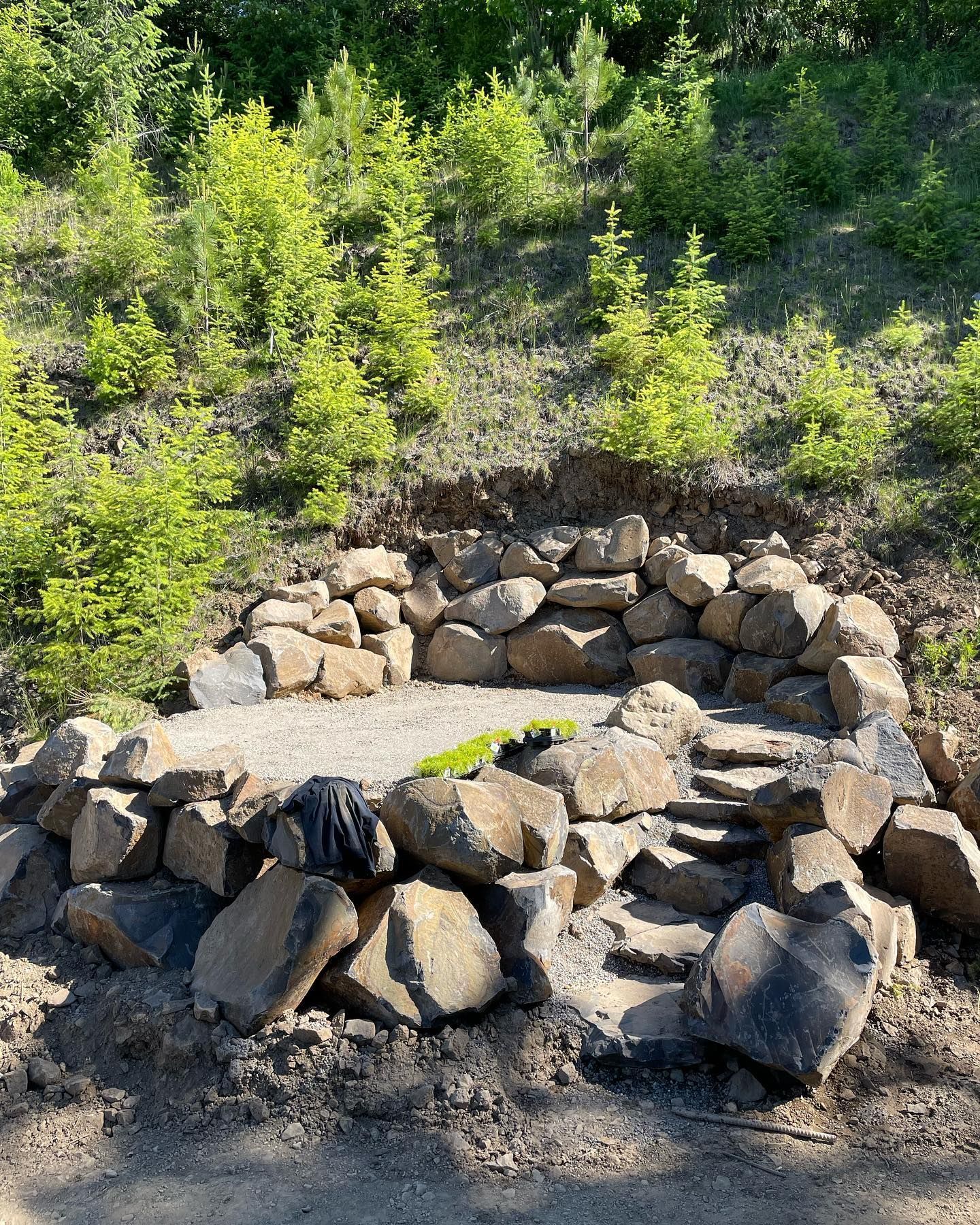 Stone-walled seating area with small steps on a hillside. Green plants and trees in background.