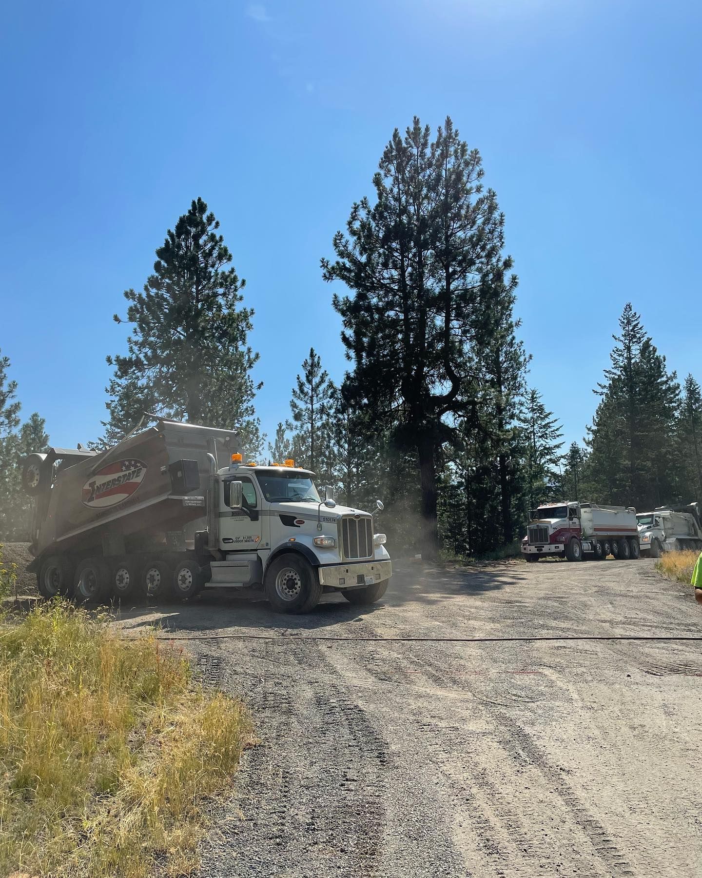 White dump truck on gravel road, with other trucks and trees in the background under a blue sky.