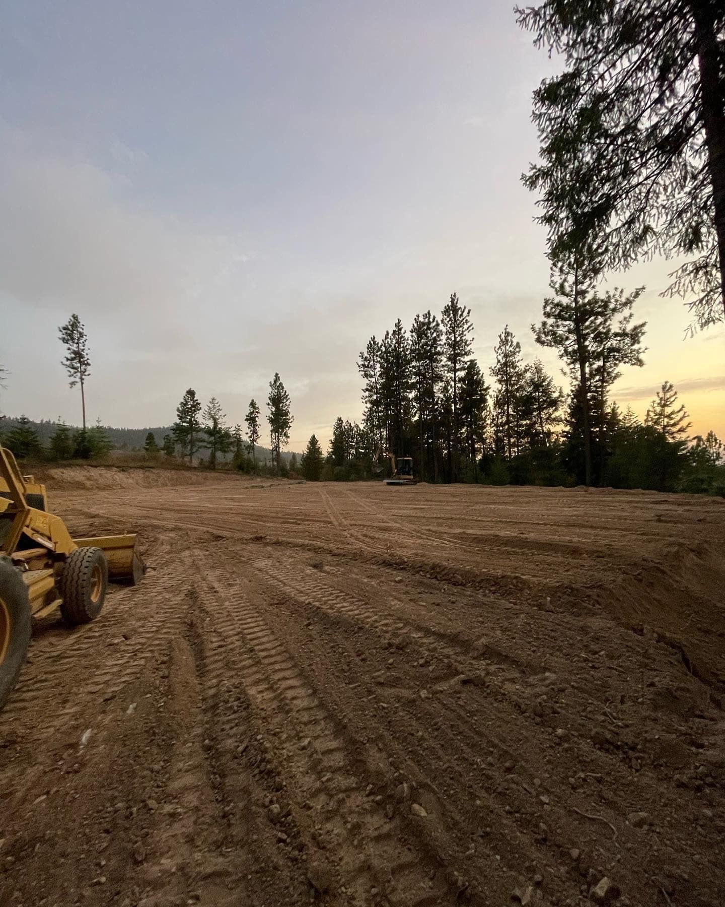 Dirt clearing with tire tracks, trees in the background, and a grading machine. Overcast sky.