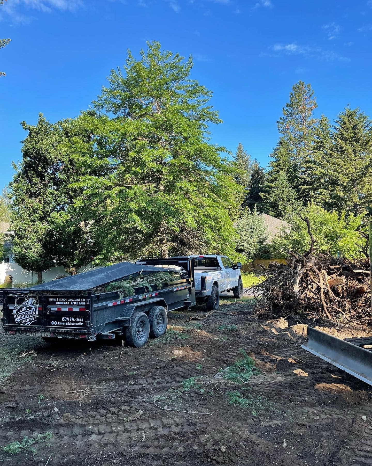 Truck pulling trailer loaded with debris on a cleared lot. Trees and a blue sky in the background.