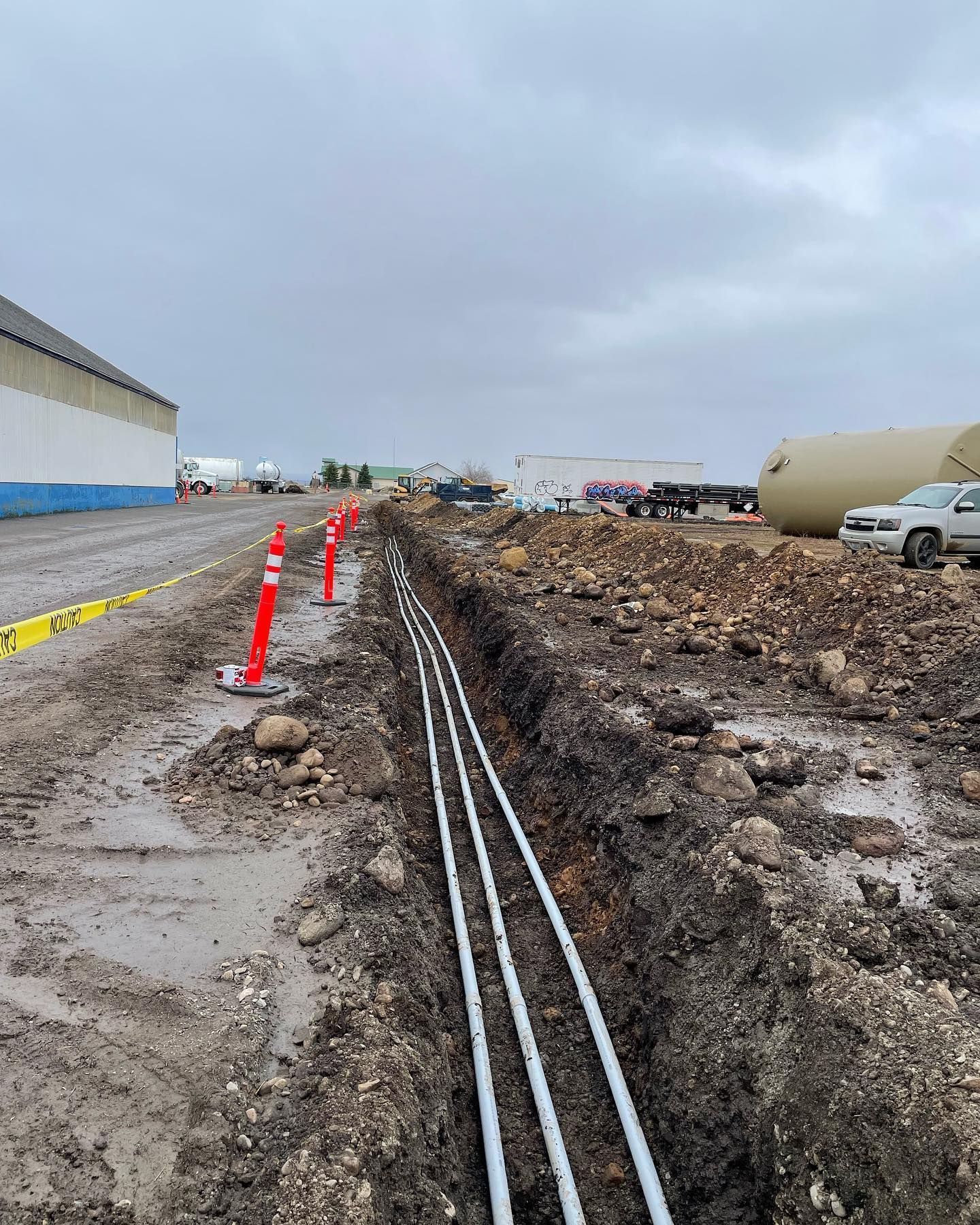 Trench with white pipes laid out, orange safety posts, and a construction site on a cloudy day.