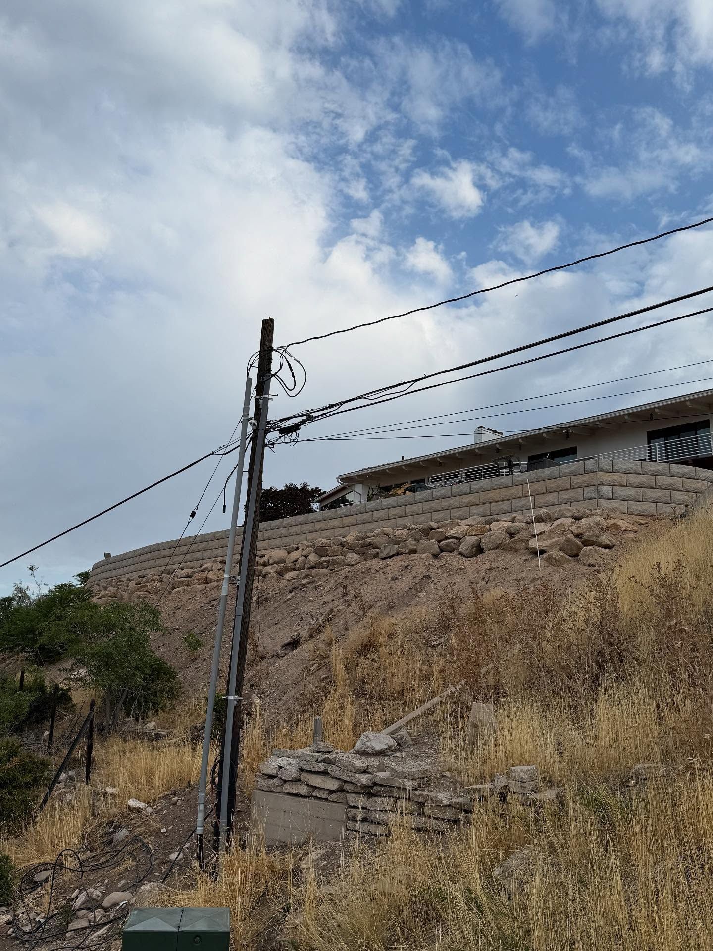 Power lines and utility pole on hillside with retaining walls and house under a partly cloudy sky.