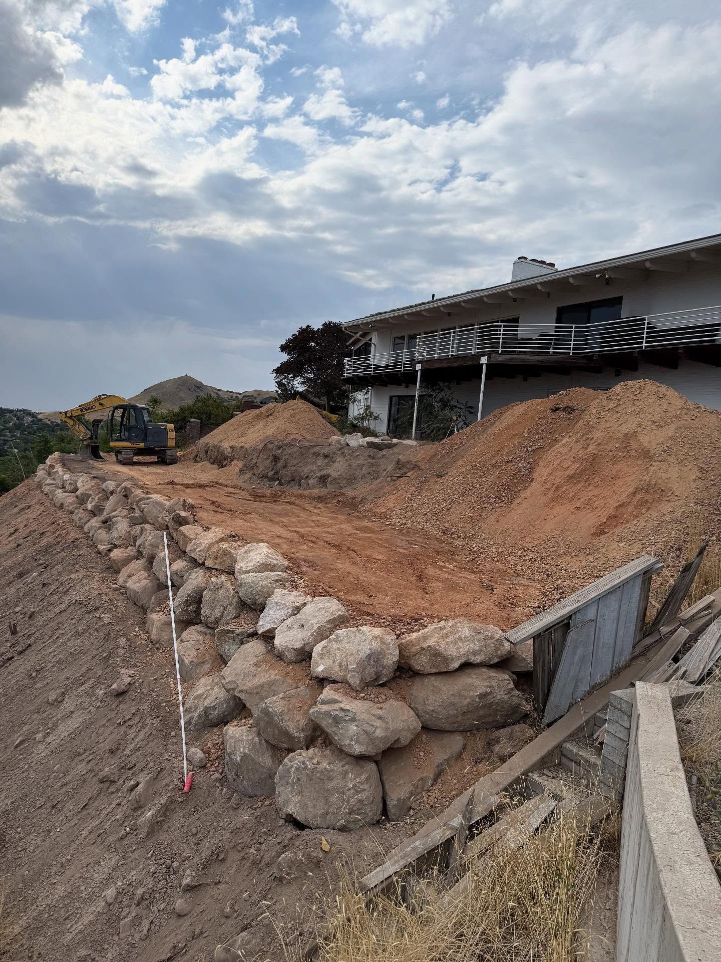 Construction site with a stone retaining wall in front of a house, cloudy sky.
