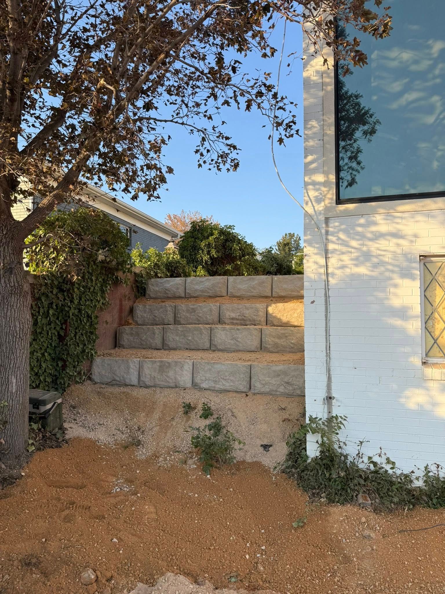 Stone steps leading uphill next to a white brick building with a large window; a tree and bushes are on the left.