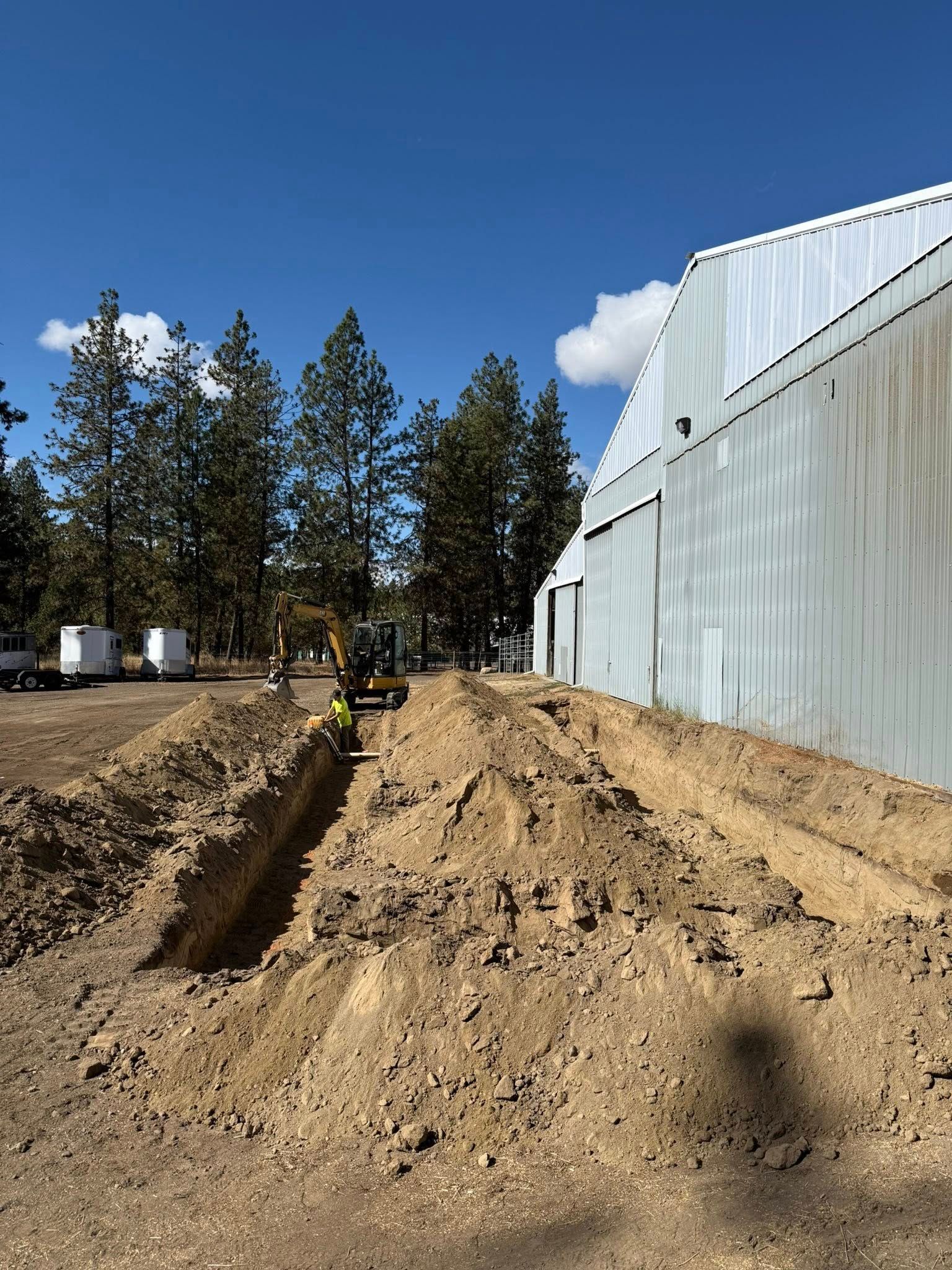 Construction site: trench being dug next to a large building.  Excavator and worker present. Blue sky.
