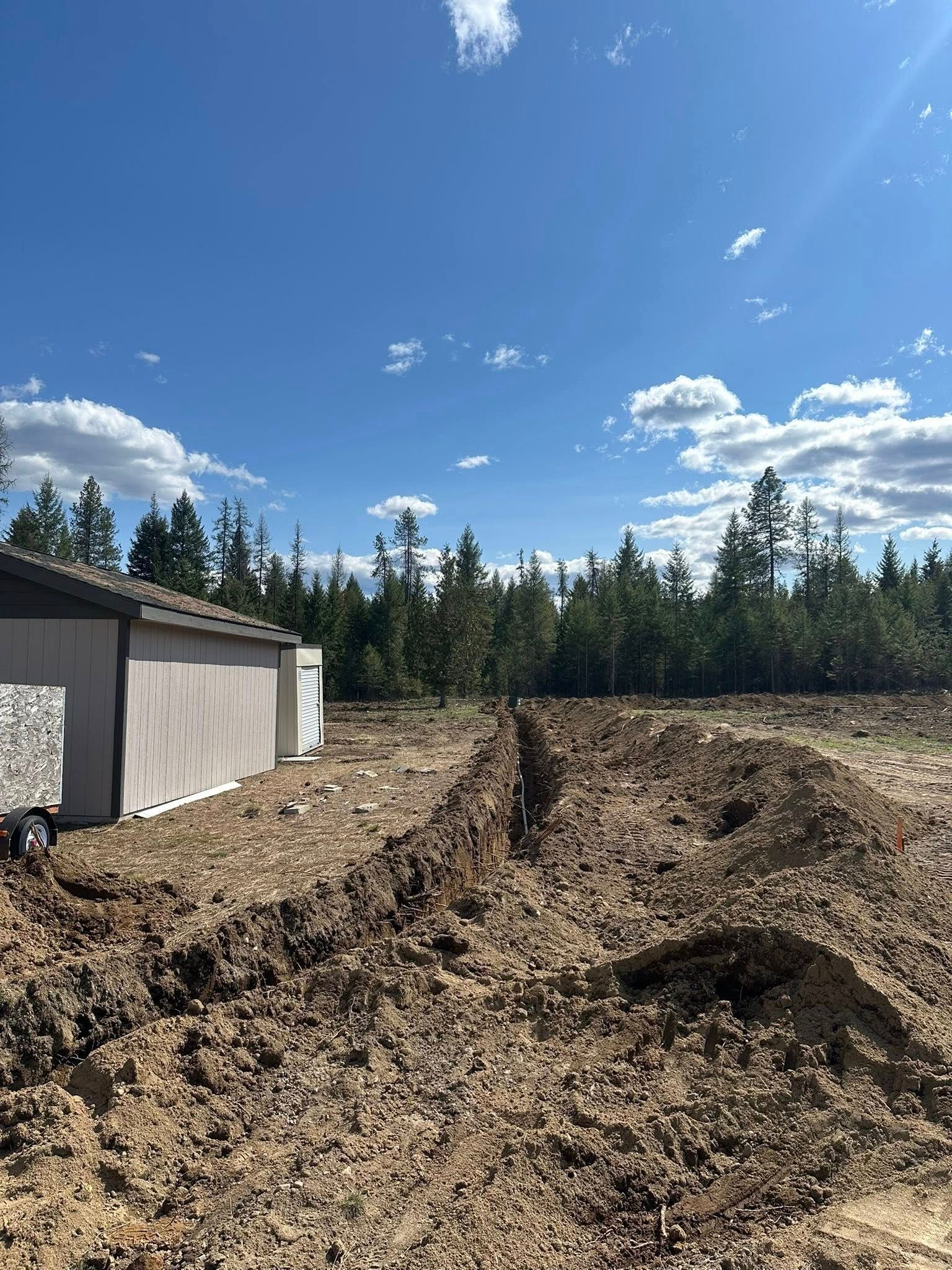 Trench dug in a dirt field next to a shed and a treeline under a blue sky with clouds.