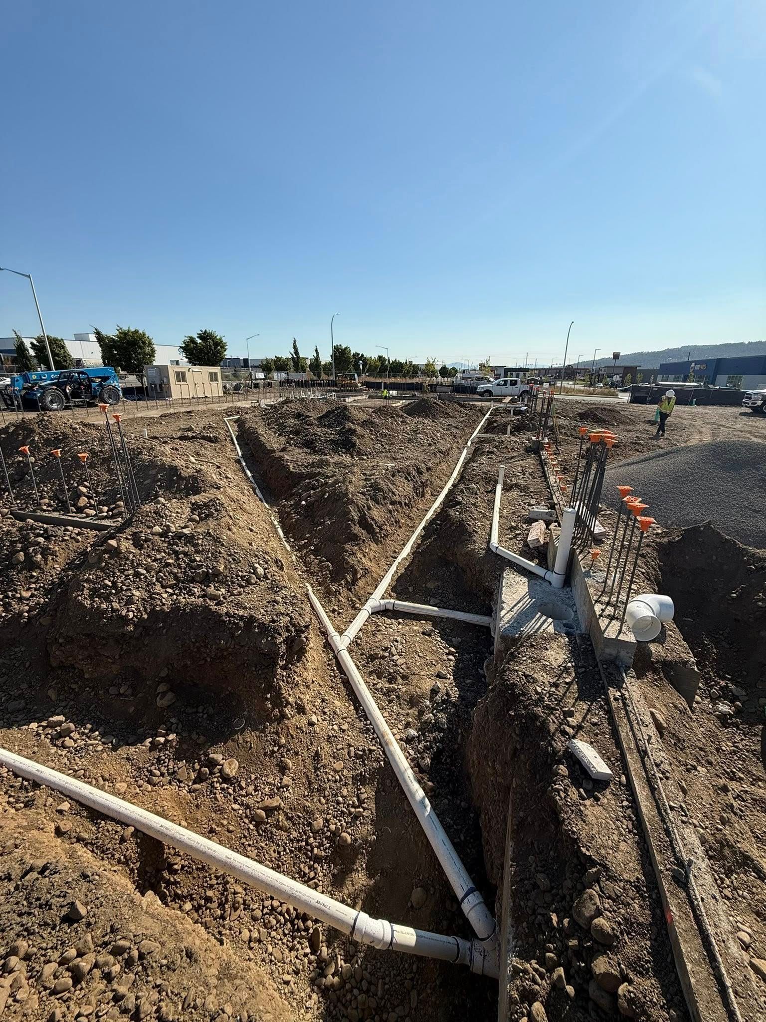 Trenches in dirt reveal white PVC pipes at a construction site on a sunny day.