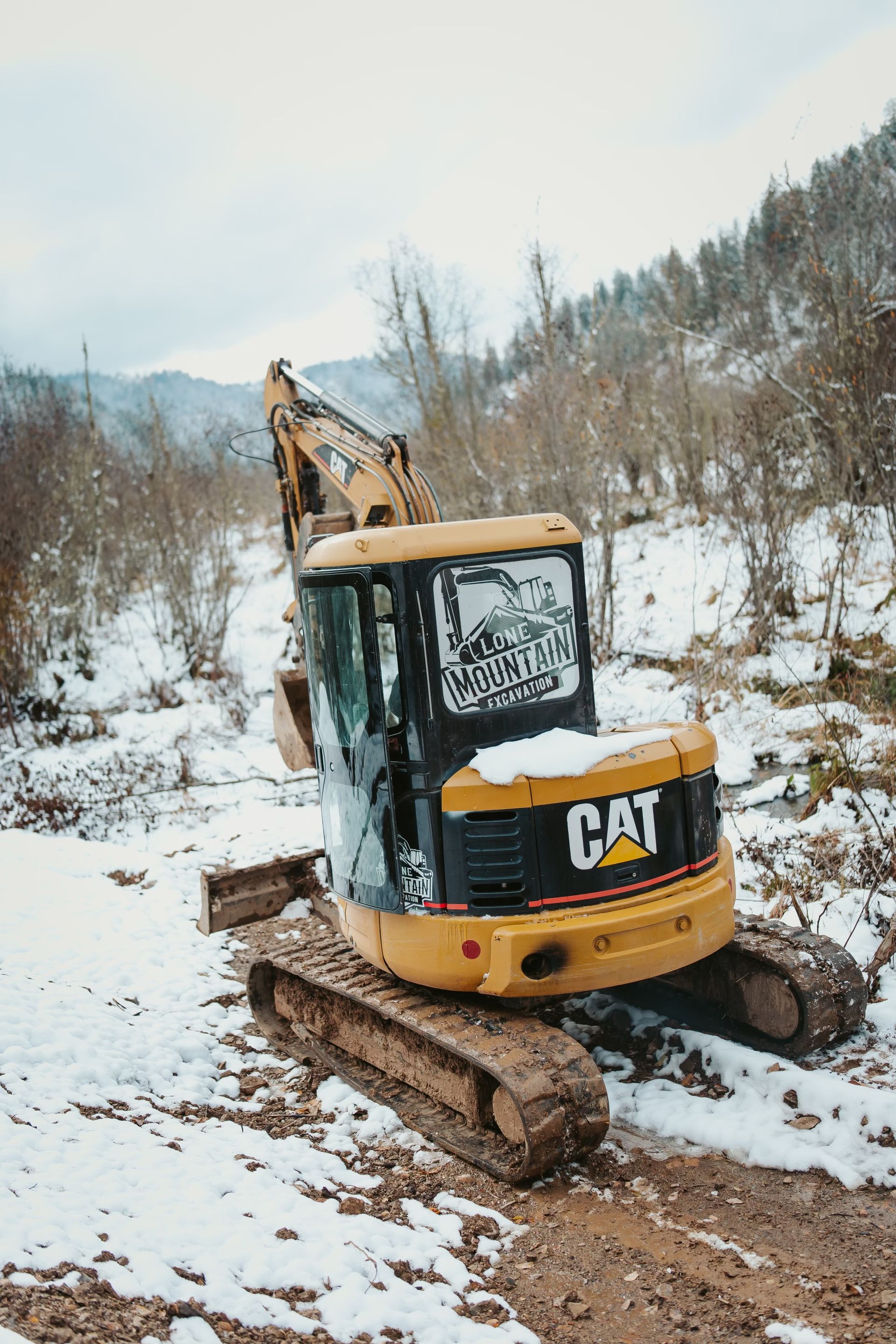 Yellow Caterpillar excavator on snowy ground. Trees and mountains in the background.
