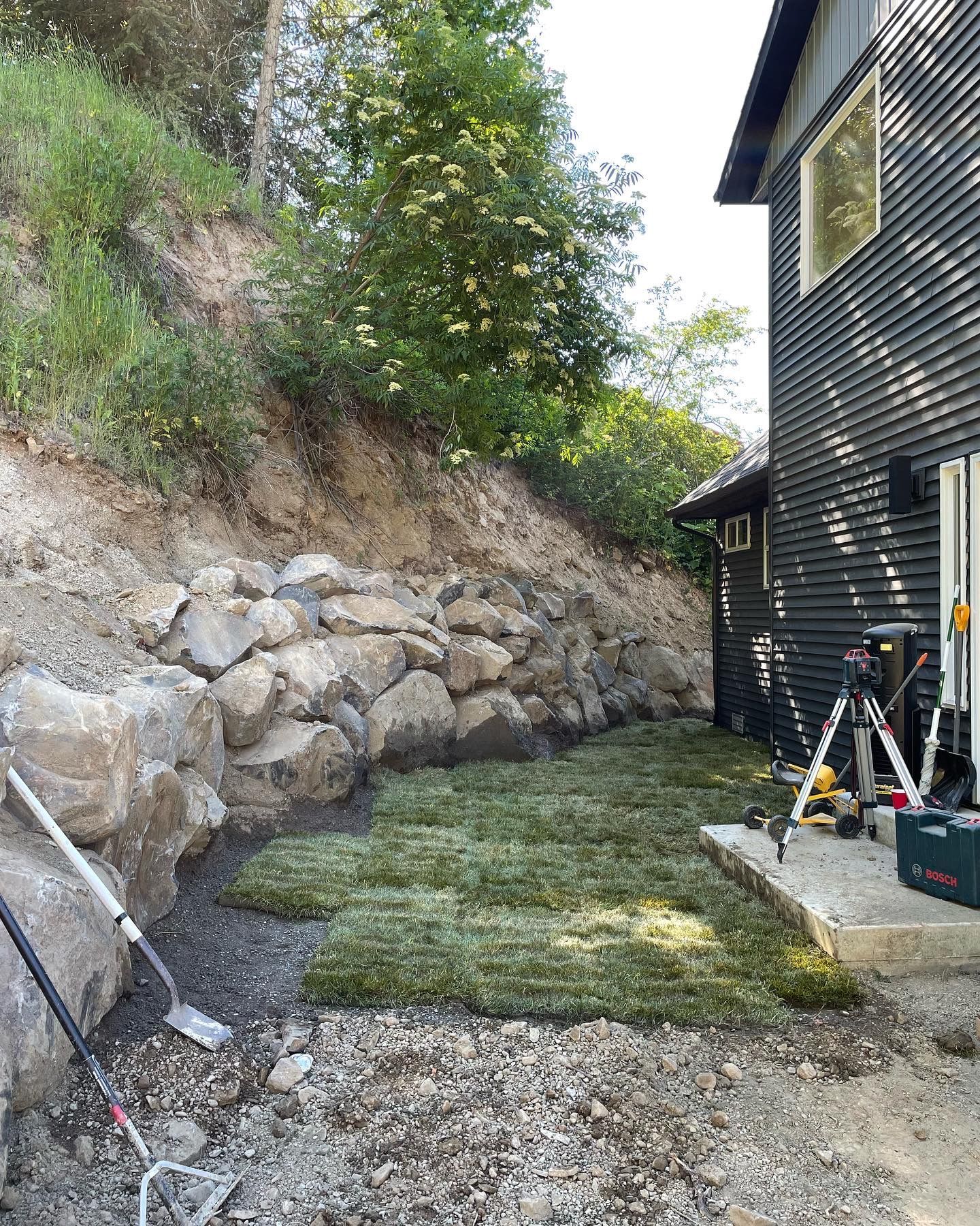 Landscaped yard with a stone retaining wall, new sod, and a dark-colored house. Construction tools are present.