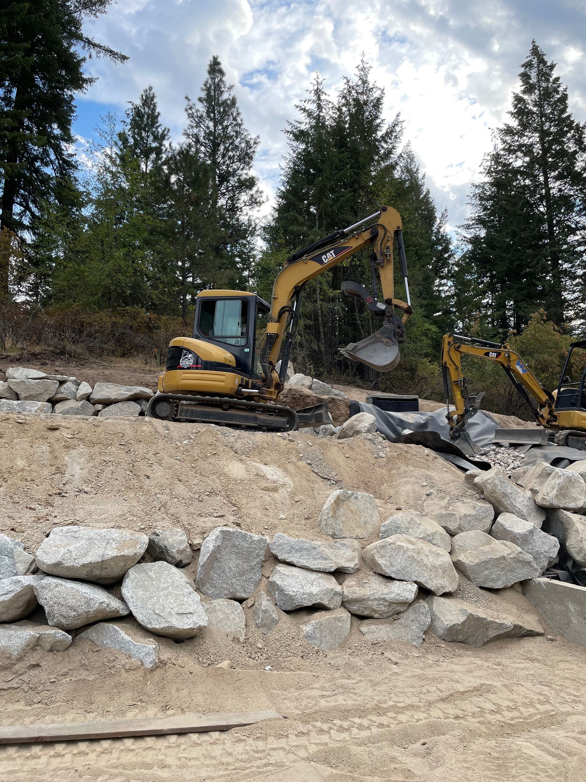 Yellow excavator on a rocky hillside, with trees in the background, working on construction.
