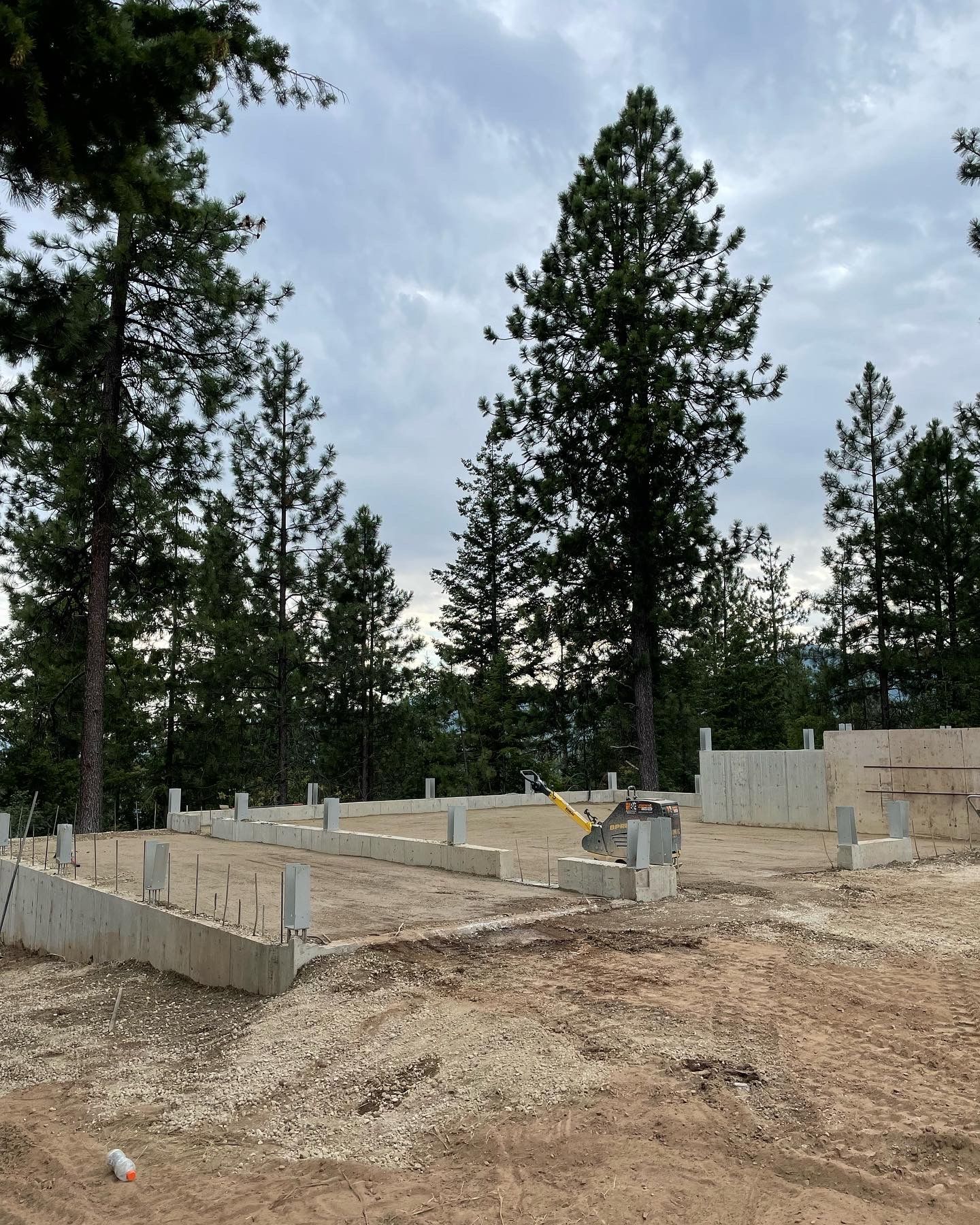 Foundation of a building under construction on a dirt lot, surrounded by tall trees under a cloudy sky.