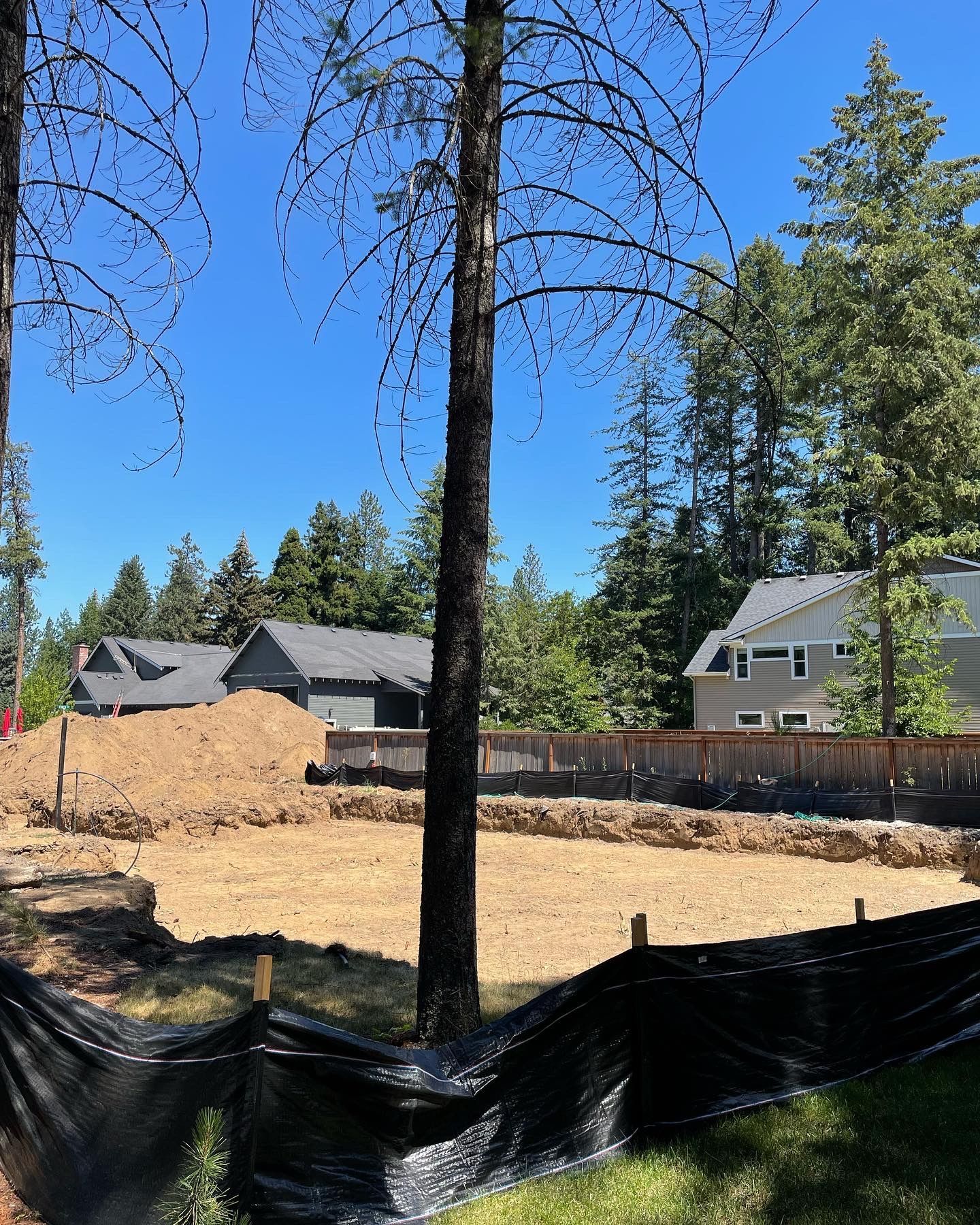 Construction site with freshly excavated lot, brown soil pile, surrounded by black fabric and trees.