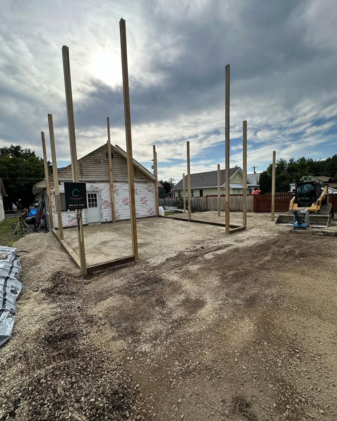 Construction site with wooden frame structure, gravel ground, small building in background. Cloudy sky.