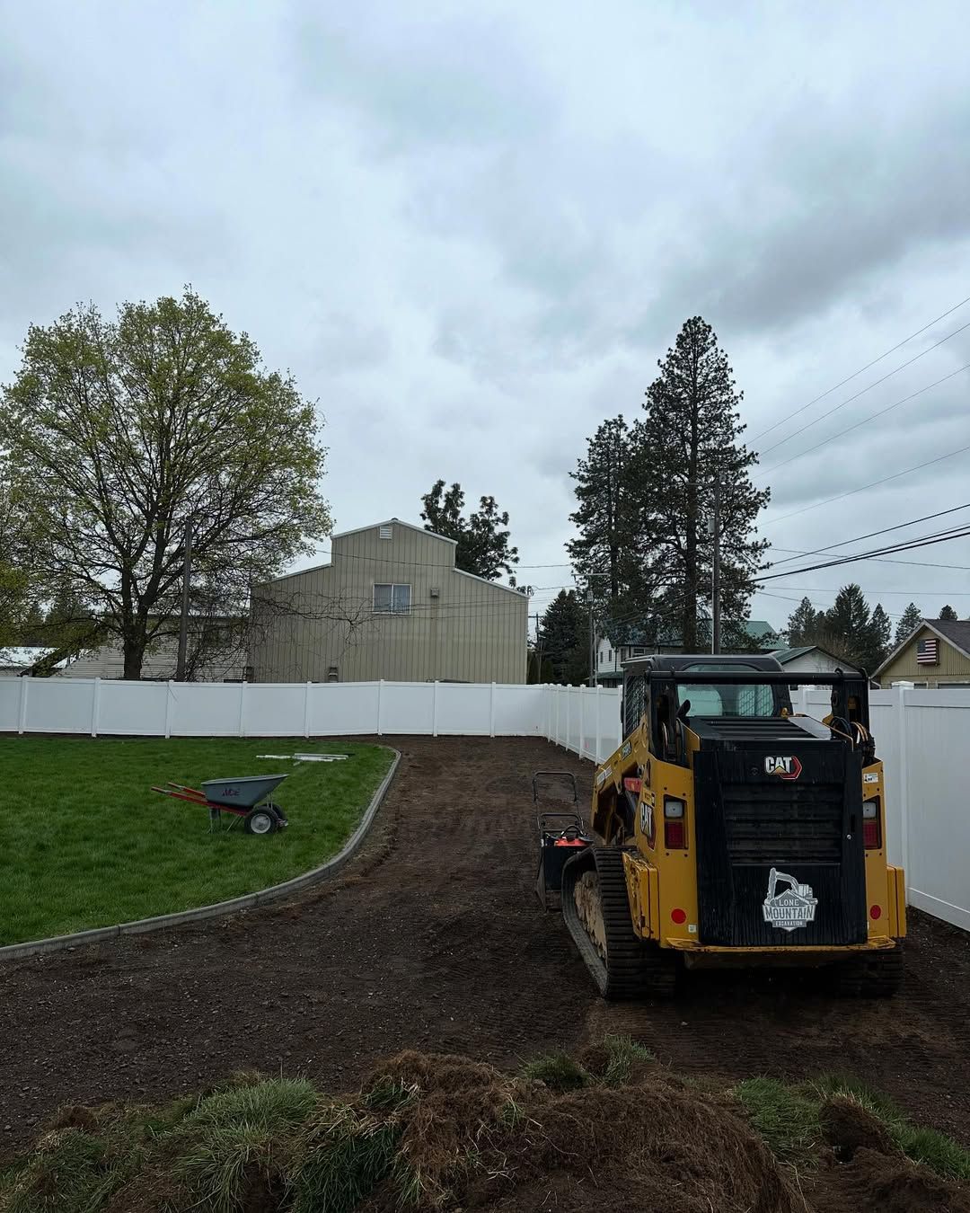 Yellow skid steer on dirt path, lawn on left. White fence surrounds. Overcast sky.